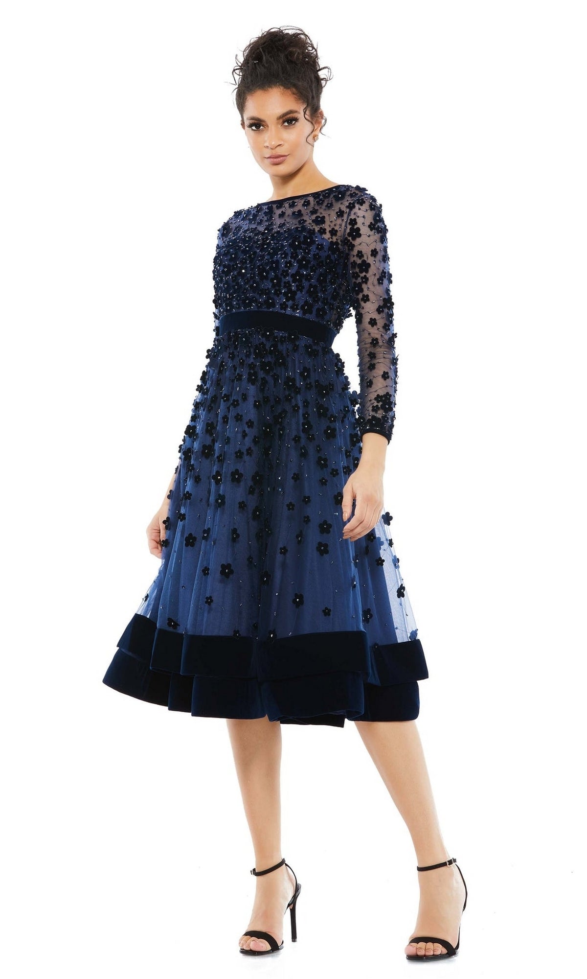 Woman in the Mac Duggal 67007 knee-length wedding-guest dress—navy blue with floral embellishments and sheer long sleeves—paired with black heels, stands against a white background; an elegant choice for any stylish guest.